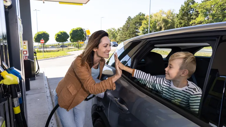 Smiling woman giving a high five to a young boy sitting in a car while refueling at a gas station.
